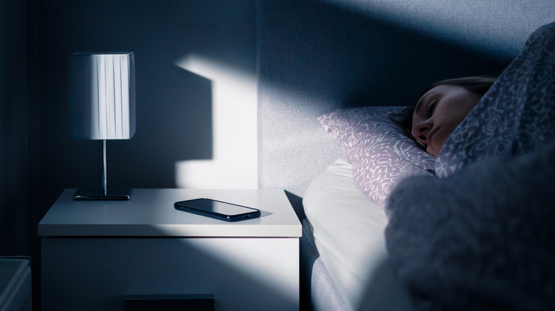 A smartphone on a nightstand next to a woman asleep in her bed