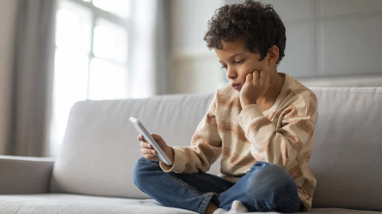 a little boy sitting on a couch and looking bored at a smartphone