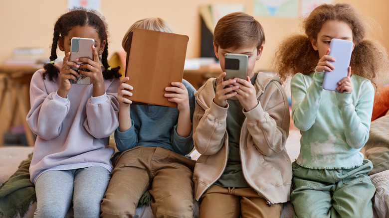 kids sitting in a row looking at smartphones and a tablet