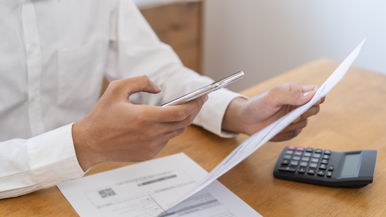person looking over paper bill while holding smartphone in hand, calculator on table