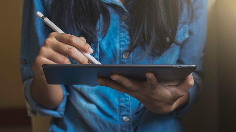 A woman in a blue button-up shirt writing on an iPad with an Apple Pencil.
