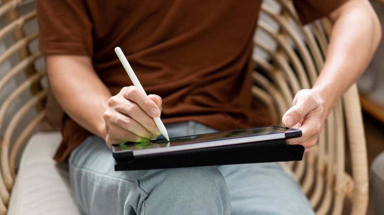 A man in a brown shirt sitting in a wicker chair using an Apple Pencil on an iPad screen