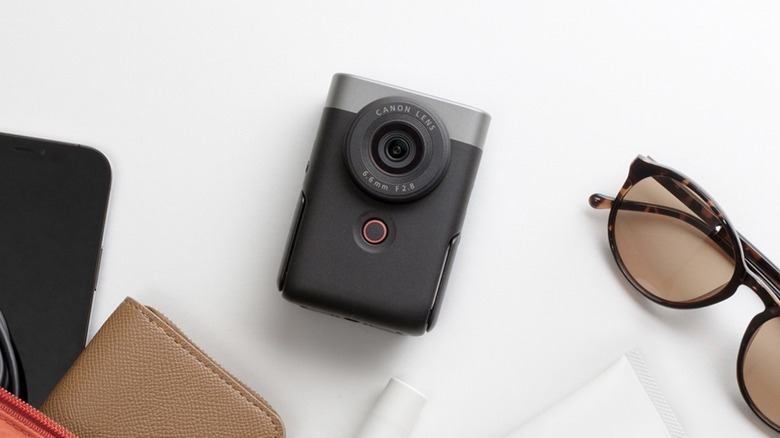 A Canon PowerShot V10 camera against a white background next to sunglasses