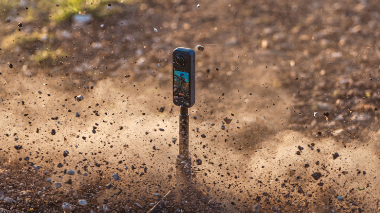 An Insta360 X5 camera on a tripod sitting on a dirt surface with debris flying all around it