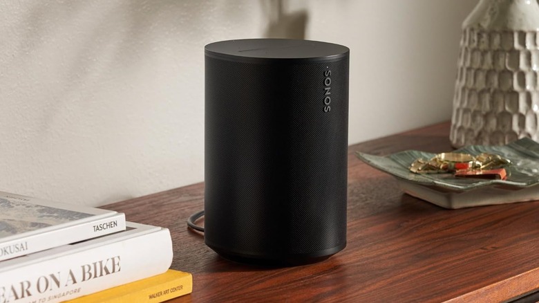 Sonos Era 100 Wireless Speaker on a table with multiple objects around it.