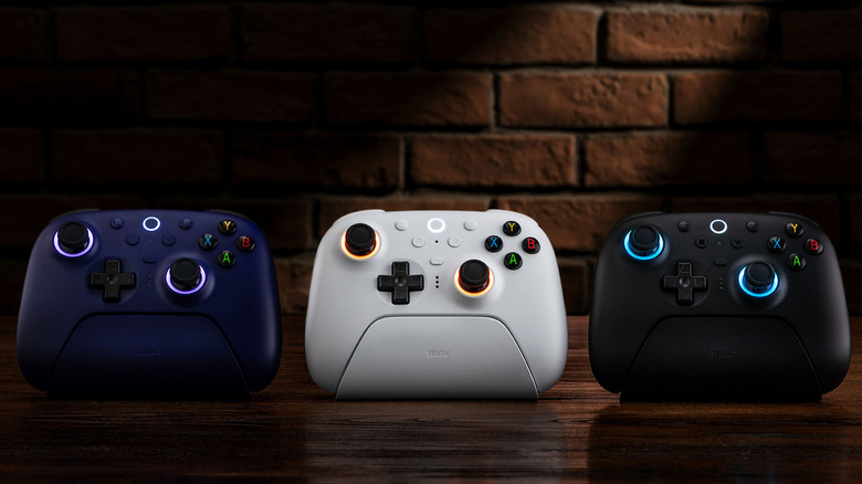 Blue, white, and black 8BitDo Ultimate 2 controllers on their charging docks, placed on wooden surface in front of a brick wall