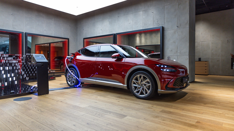 A red 2023 Genesis GV60 parked in a lit garage display, plugged into a charger.