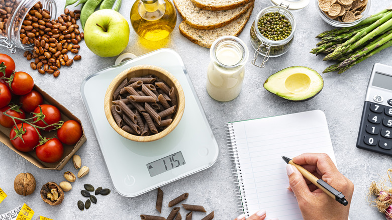 A food scale surrounded by healthy foods, and a person's hand holding a pen over a notebook.