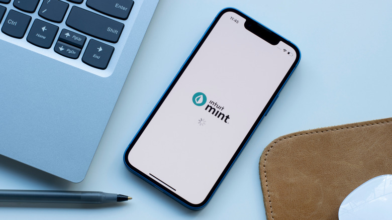 The Mint app loading screen on an iphone that's sitting on a table next to a notebook and a laptop