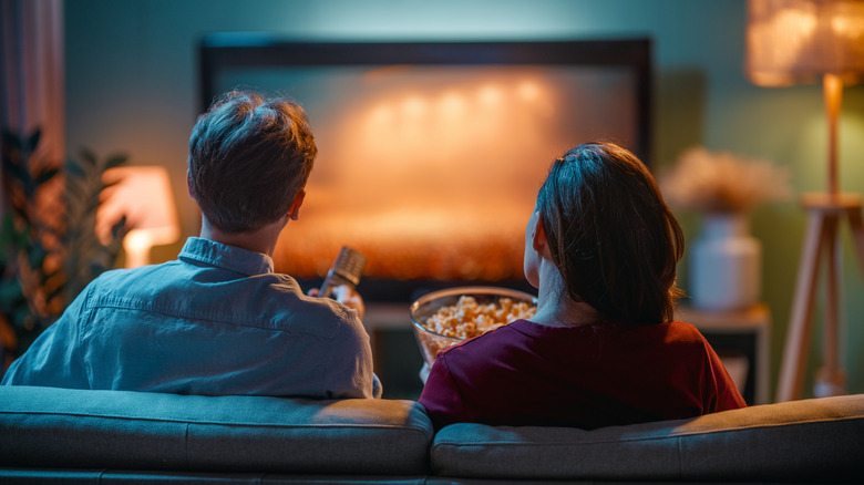 Couple sitting on a couch with popcorn while watching TV in a warmly lit living room.
