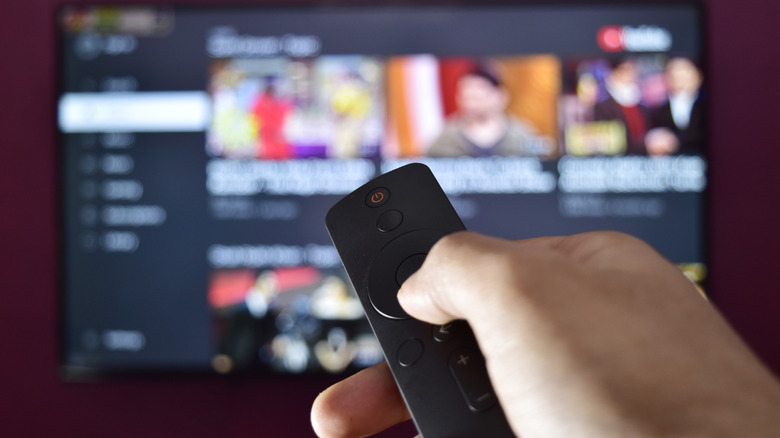 Hand pressing a button on a streaming remote in front of a blurred TV app menu.