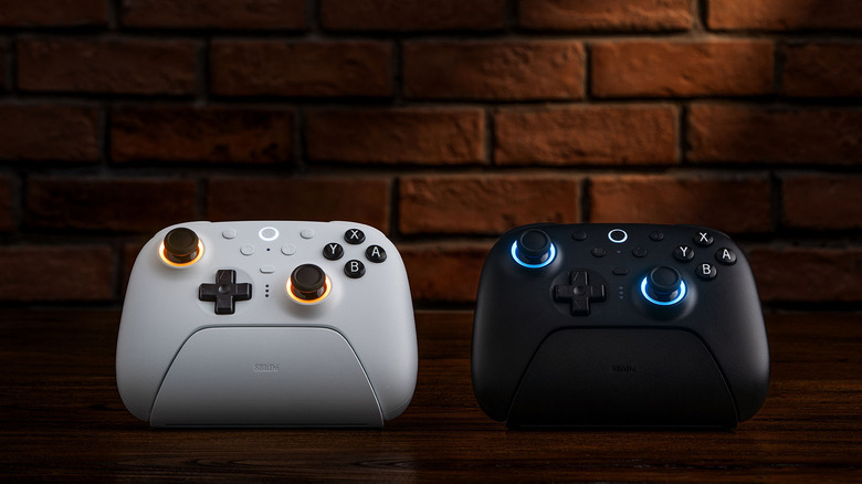 Two 8BitDo Ultimate 2 controllers, white and black, side by side in matching docks in front of a brick wall