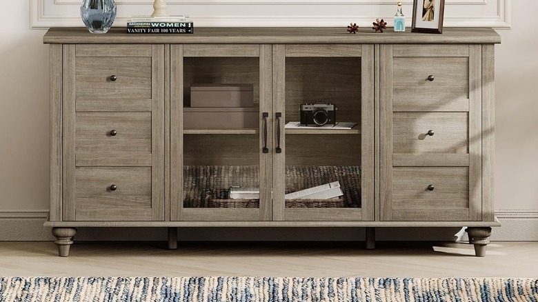 The Wampat Farmhouse TV Stand set up in a living room. Various items adorn the top of the stand, and there are several items in its middle compartment.