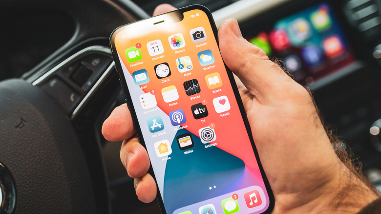A closeup of an iPhone on the Home screen in front of a vehicle displaying CarPlay on its infotainment system