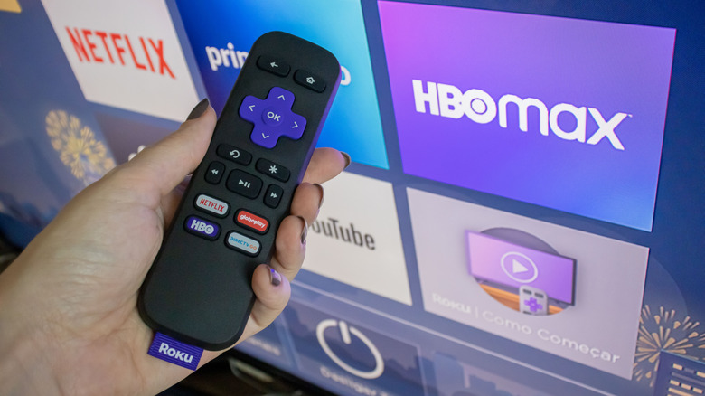 A hand holding a Roku remote in front of a TV displaying several streaming apps within Roku OS