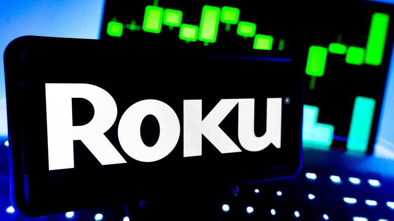 A smartphone displaying the Roku logo, in the background is a screen displaying green and teal graphs