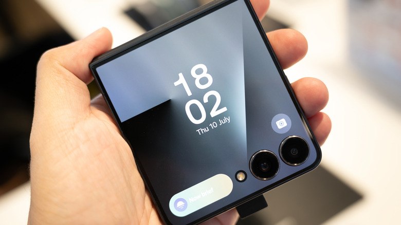 A person holding the Samsung Galaxy Z Flip7 in their hand with the time displayed on the cover screen