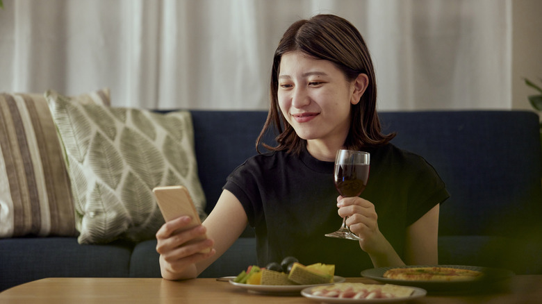 An individual sitting on the floor in between a couch and a table holds a glass of wine in one hand a smartphone in the other, the person has three plates of food in front of them on the table