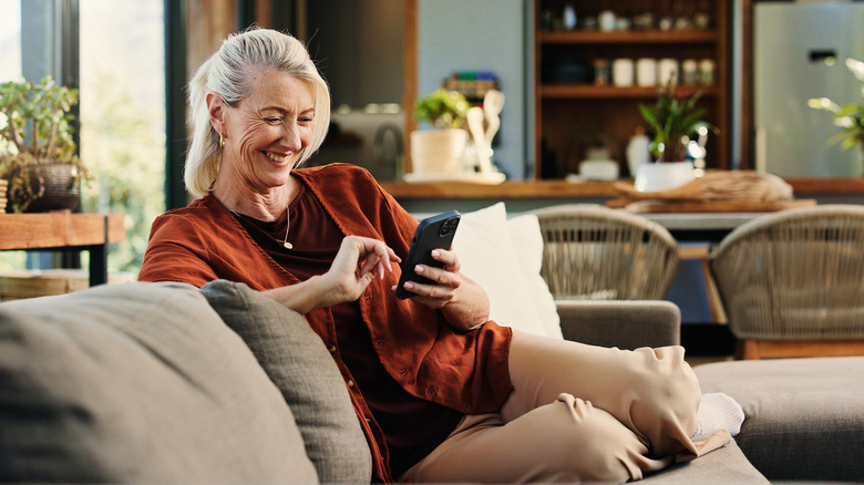 An individual with silver-blonde hair and a red shirt sits on a couch looking at their black cellphone