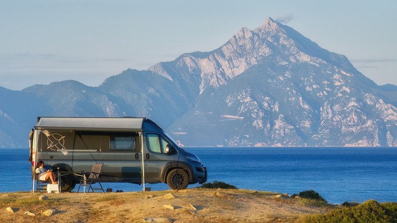 A person sits outside of a parked camper van with a mountain vista in the background.