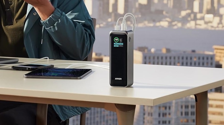 The Anker Prime Power Bank 27,650 mAh is placed on a table in an outdoor setting. A person sits next to it, and there's both a phone and a tablet connected for charging.
