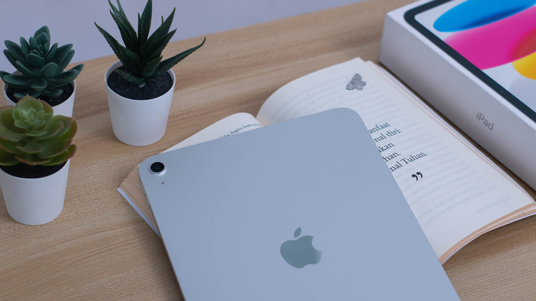 A silver iPad lays on a book that's on a desk, next to the iPad and book is the iPad's original box, there are also three small potted plants in the image