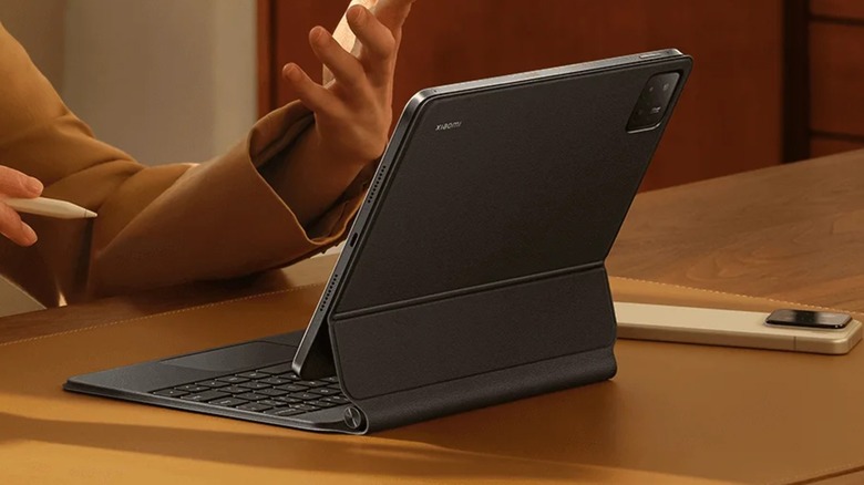 A black Xiaomi Pad 7 with a keyboard folio case sits next to a white smartphone on top of a desk, an individual holding a tablet pen can be seen cropped from the photo