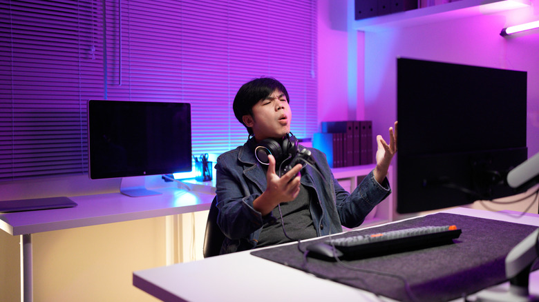 A man looking at his computer screen with headphones around his neck, making a frustrated expression and holding his hands up in disbelief