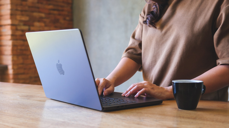 A person using a MacBook as it sits on a wooden table, a coffee cup is next to the individual's left hand