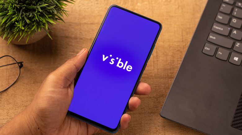 A hand holding a phone showing a splash logo screen of the Visible cell phone carrier.