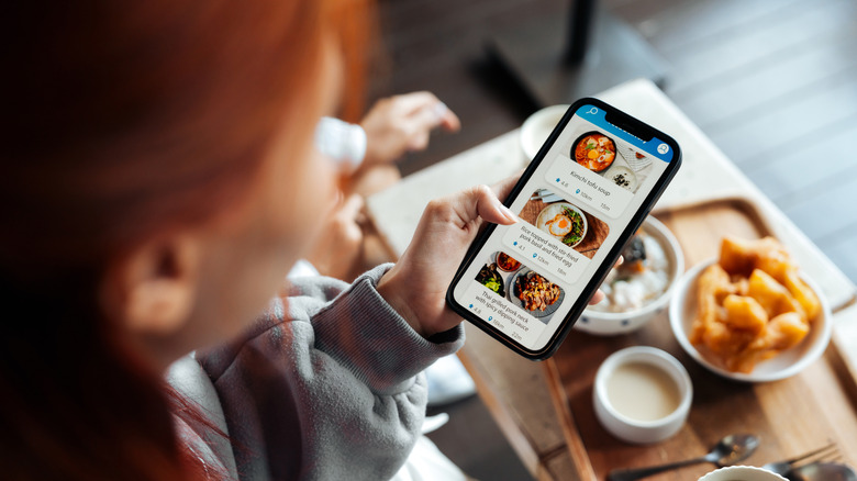 A woman holding an iPhone with a food app open and a food tray in the background.
