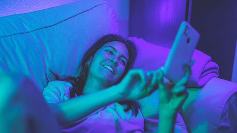 A woman lying on a sofa smiling at her phone under cyan and purple lights.