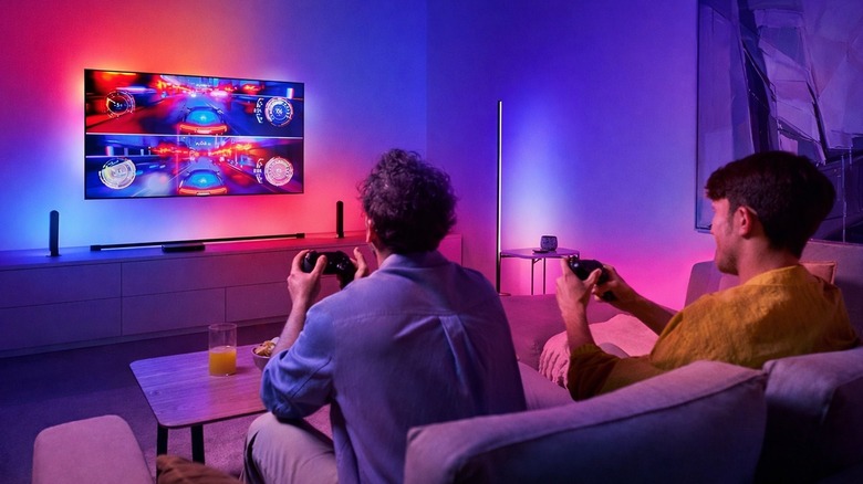 Two men on a couch playing a racing game on a TV with red-purple background lighting.