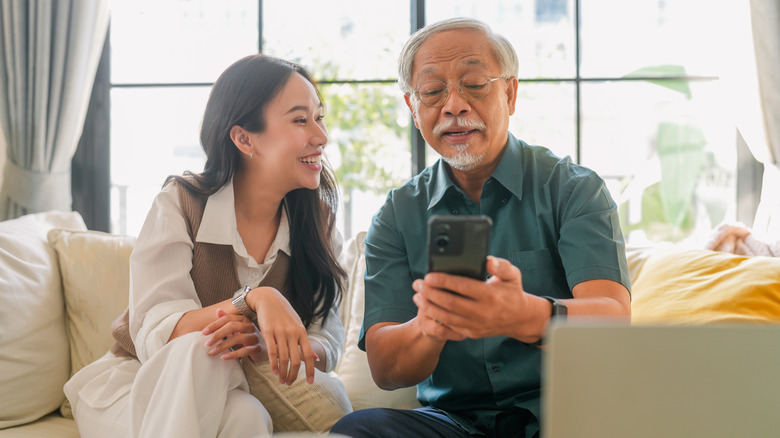 An elderly man uses a smartphone with a young woman smiling beside him.