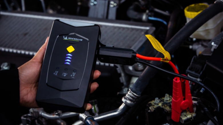 The Michelin Portable Jump Starter being held in an individual's left hand as it is connected to a car battery, presumably to give the battery a jump