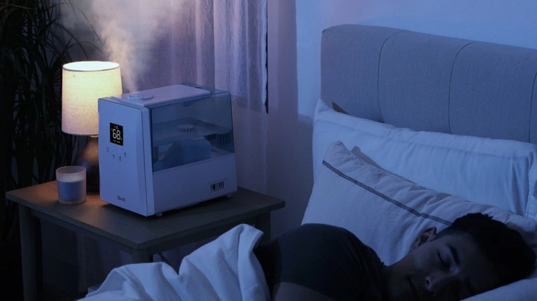 The Levoit LV600S humidifier on a bedside table next to a man sleeping in a bed.