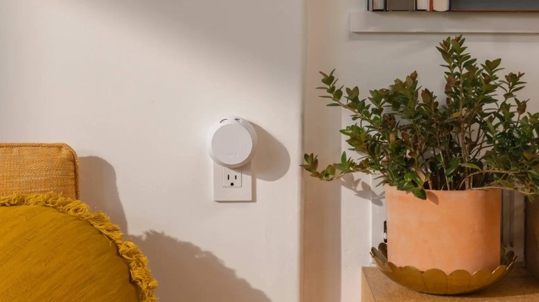The Pura 4 smart fragrance diffuser plugged into a wall outlet, beside a plant and a chair.