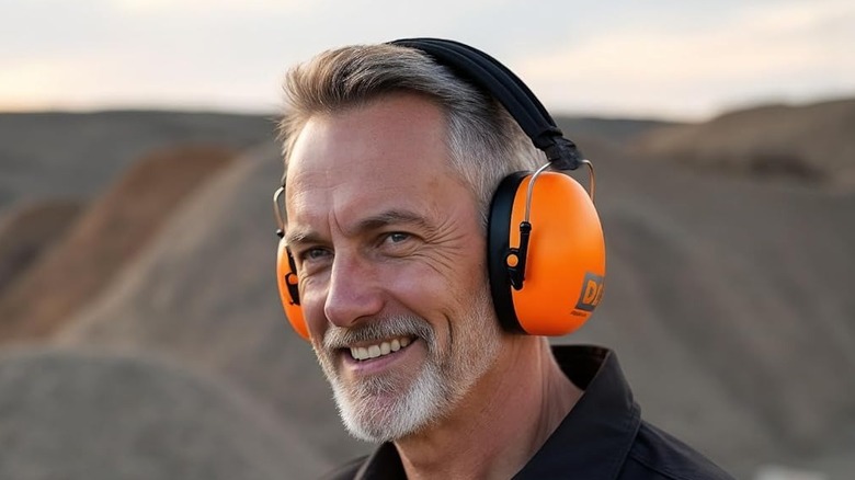 A smiling man wearing orange Decibel Defense Safety Ear Muffs with blurred mountains in the background.