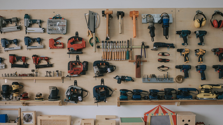 Several hand tools and power tools mounted on a wooden board on a gray wall.