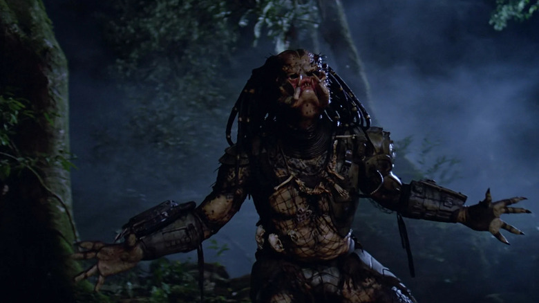 The Predator screaming with his hands open wide in the dark jungle