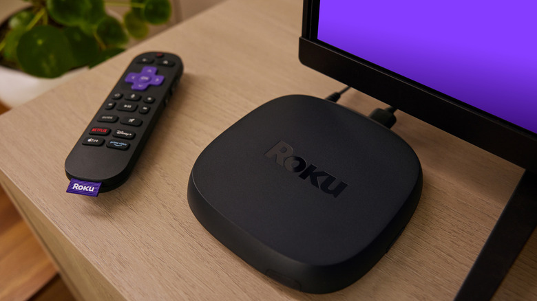 The Roku Ultra (2024) set up on top of a TV stand. Its remote is placed next to it. The corner of a TV is above it.