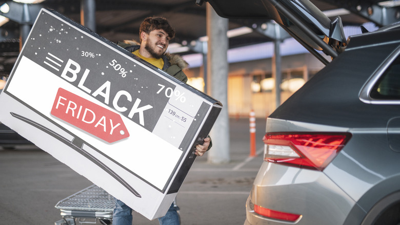 Smiling man loading large Black Friday TV box into car trunk in parking garage.
