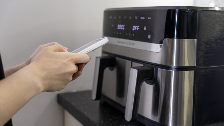 Hands using an app on a smartphone to control an air fryer on a kitchen counter