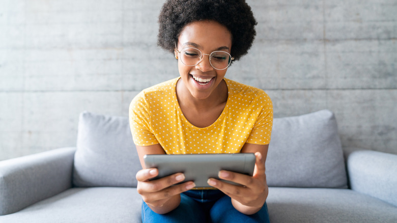 a woman smiling and looking at her tablet while sitting on a gray sofa