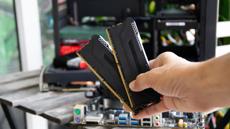 A hand holding two DDR5 RAM sticks in front of a blurred open desktop computer build