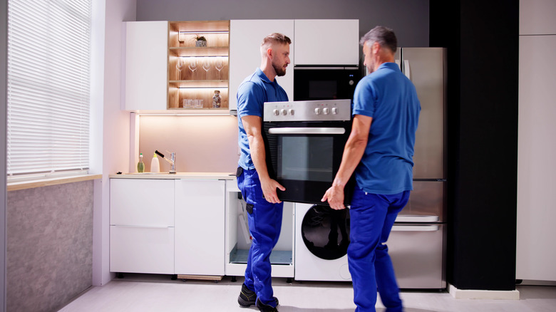 Two individuals dressed in blue shirts and pants are carrying an oven as they stand in a mostly white kitchen