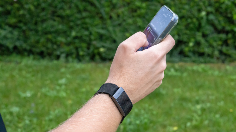 A black Whoop band on someone's wrist while using their smartphone in a grassy area.