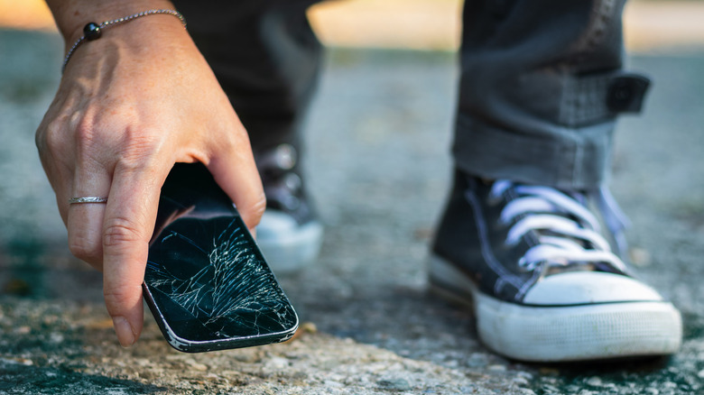 A man's hand retrieving a broken phone from the ground.
