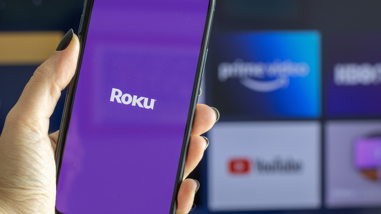 Roku logo on a smartphone as the Roku app loads, TV display blurred in background