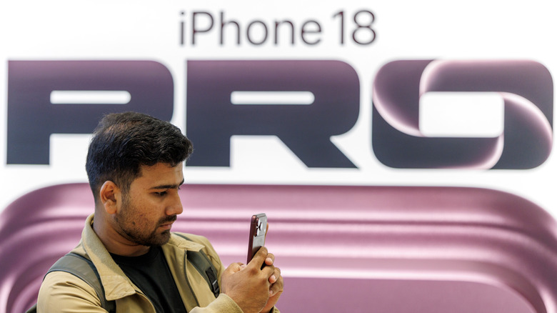A person using an iPhone 17 Pro Max in front of an iPhone 18 Pro billboard.